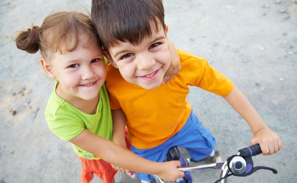 Ein kleines lächelndes Mädchen umarmt einen kleinen Bub, der auf ein Fahrrad sitzt